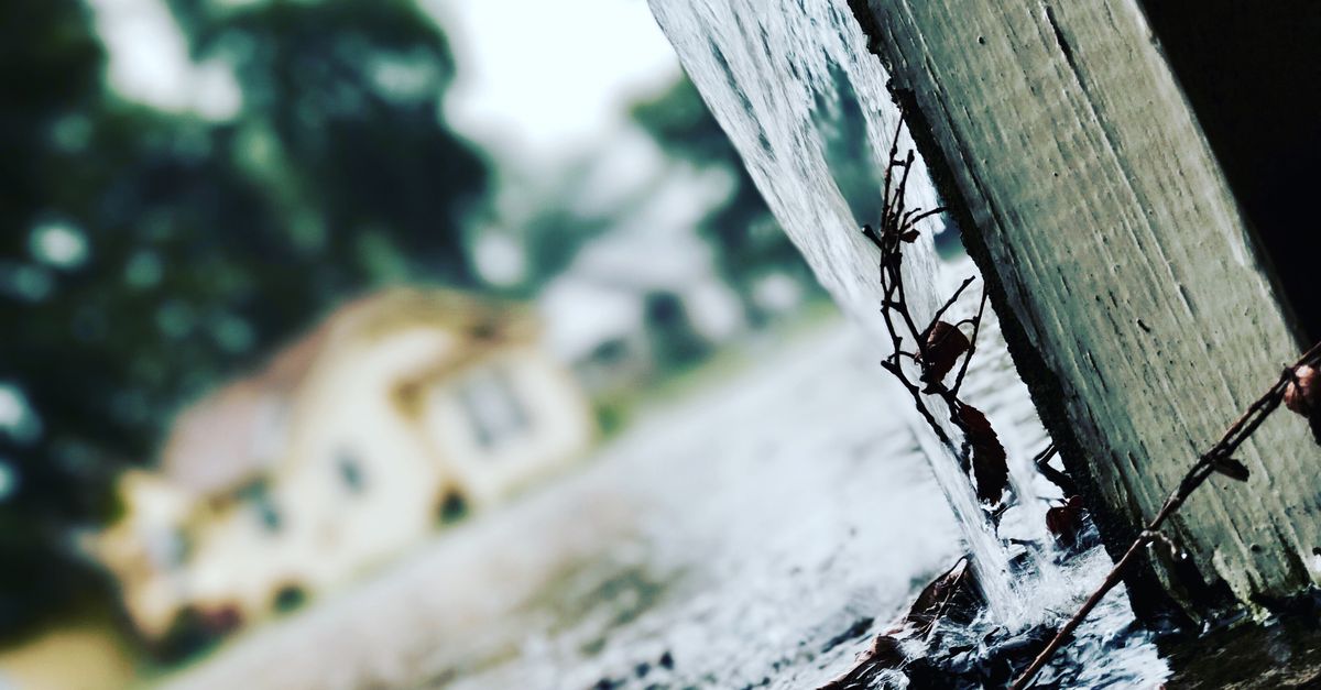 Rainwater flowing from a metal gutter downspout during a rainstorm, showing the runoff that a first flush diverter would divert before it reaches a storage tank