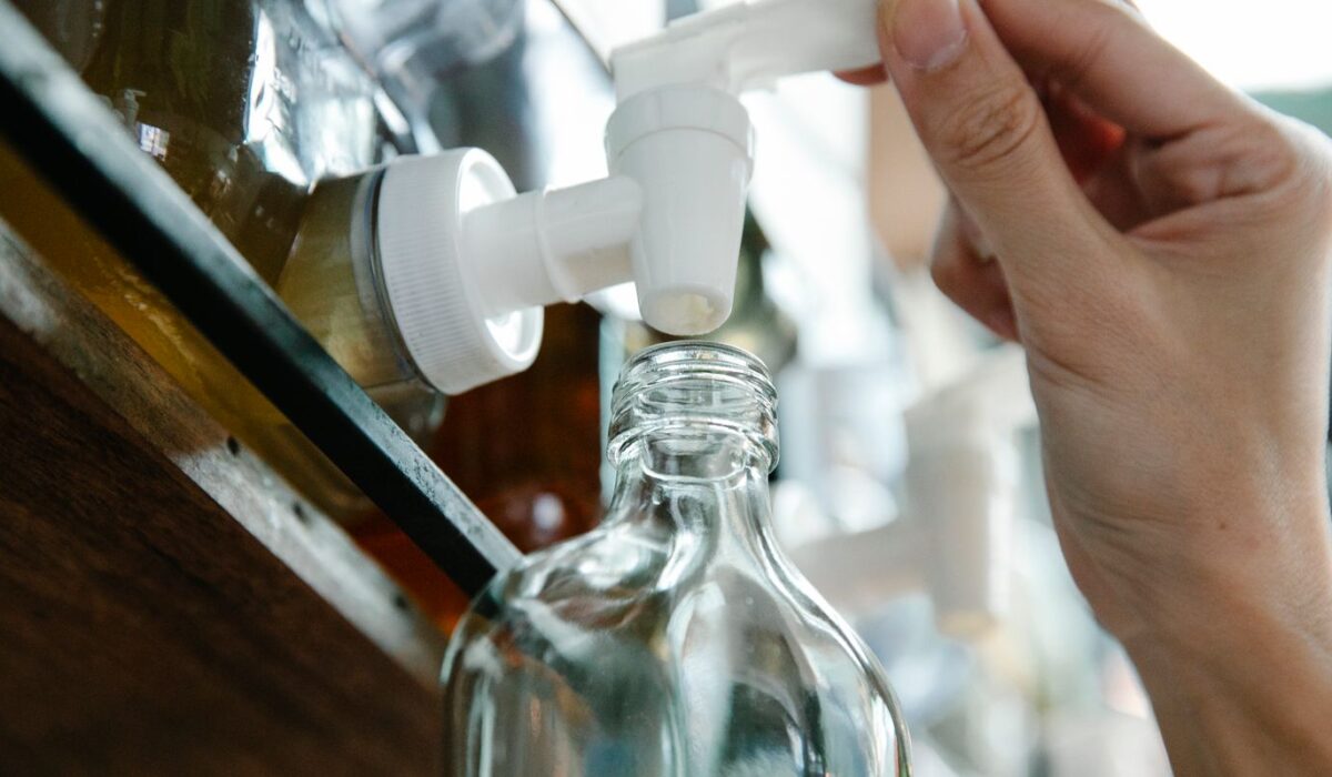 Hand refilling a glass cleaning-solution bottle from a pump dispenser on a kitchen counter.