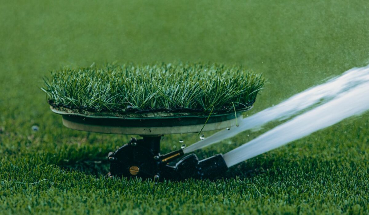An in-ground lawn sprinkler head spraying an arc of water over green turf during early morning light.