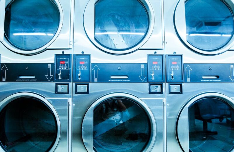 Row of modern front-loading washing machines in a bright launderette, showing cycle selector dials and door windows.